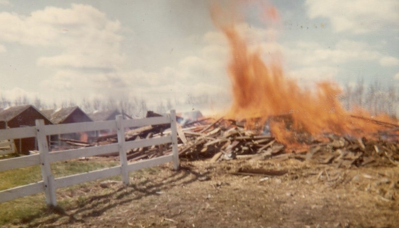 A pile of old barn boards burning in the farmyard beside a white fence, flames rising against a cloudy prairie sky